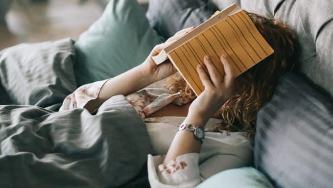 Persona tumbada en una cama con cabecero gris, cubriéndose la cara con un libro amarillo abierto, rodeada de almohadas y mantas.