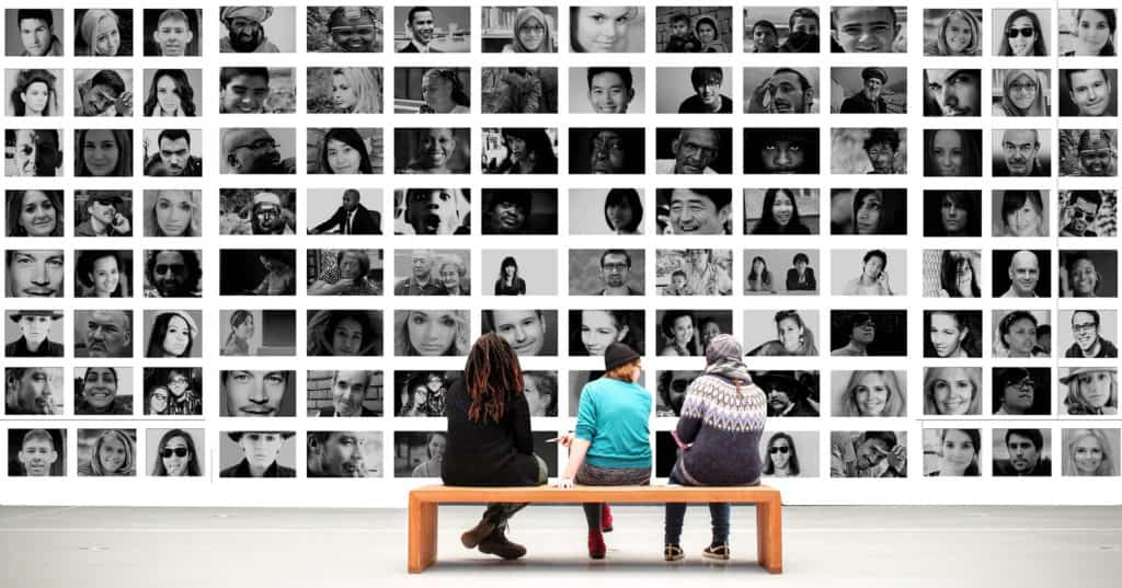 Tres personas sentadas en un banco frente a una gran pared cubierta con una cuadrícula de retratos en blanco y negro de individuos diversos.
